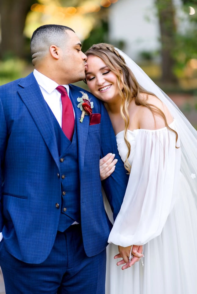 Groom in a blue suit and red tie kissing bride on the cheek, with blurred outdoor background