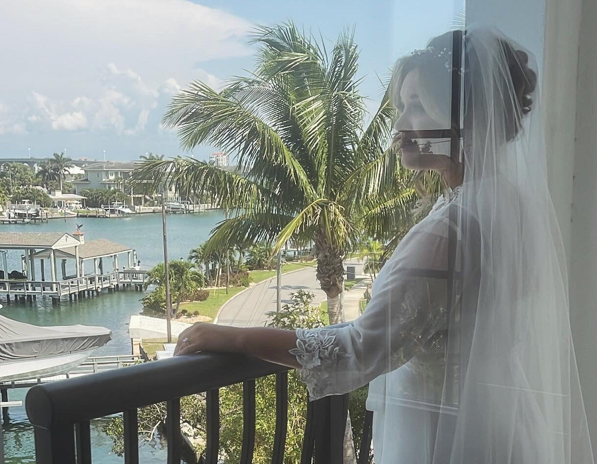 Bride in a white dress standing on a balcony with a scenic view of water and palm trees.
