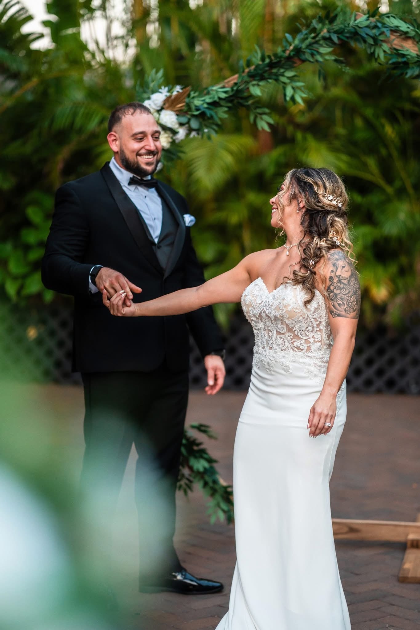 Man and woman in formal attire standing close together outdoors with greenery around.